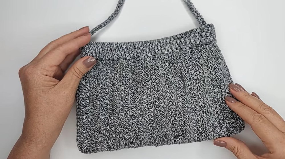 Clutch de crochê cinza sendo segurada pelas duas mãos, mostrando a estrutura e textura canelada da bolsa feita com Fio Cisne Original
