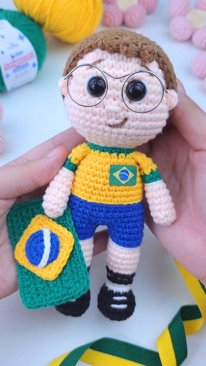 Amigurumi Garoto Torcedor segurando a bandeira do Brasil, com camisa da seleção, óculos redondos e chuteiras pretas