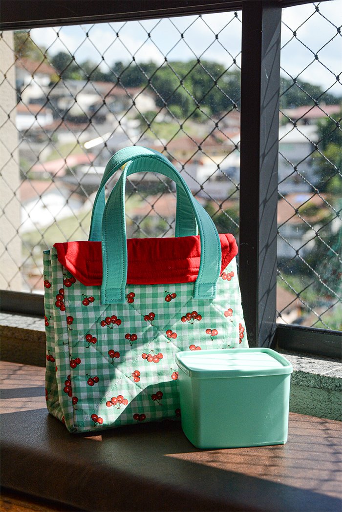 Lancheira de Cerejas em patchwork ao lado de um pote verde, mostrando o tamanho ideal para levar o lanche