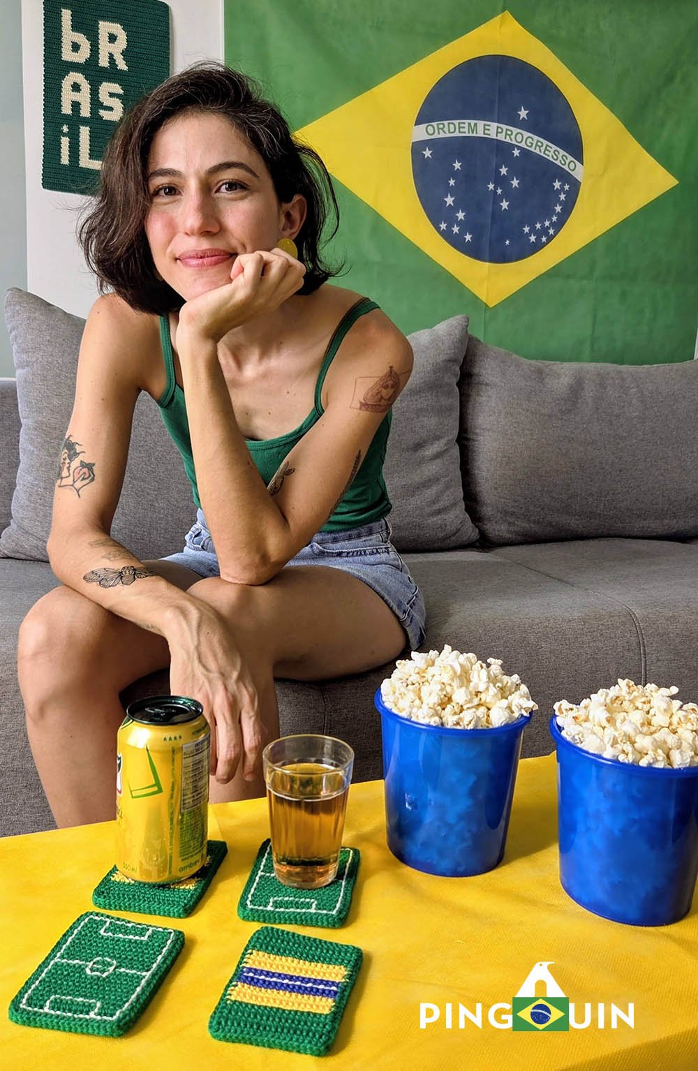 Artesã com porta-copos de crochê temáticos do Brasil sobre mesa amarela com bandeira brasileira ao fundo