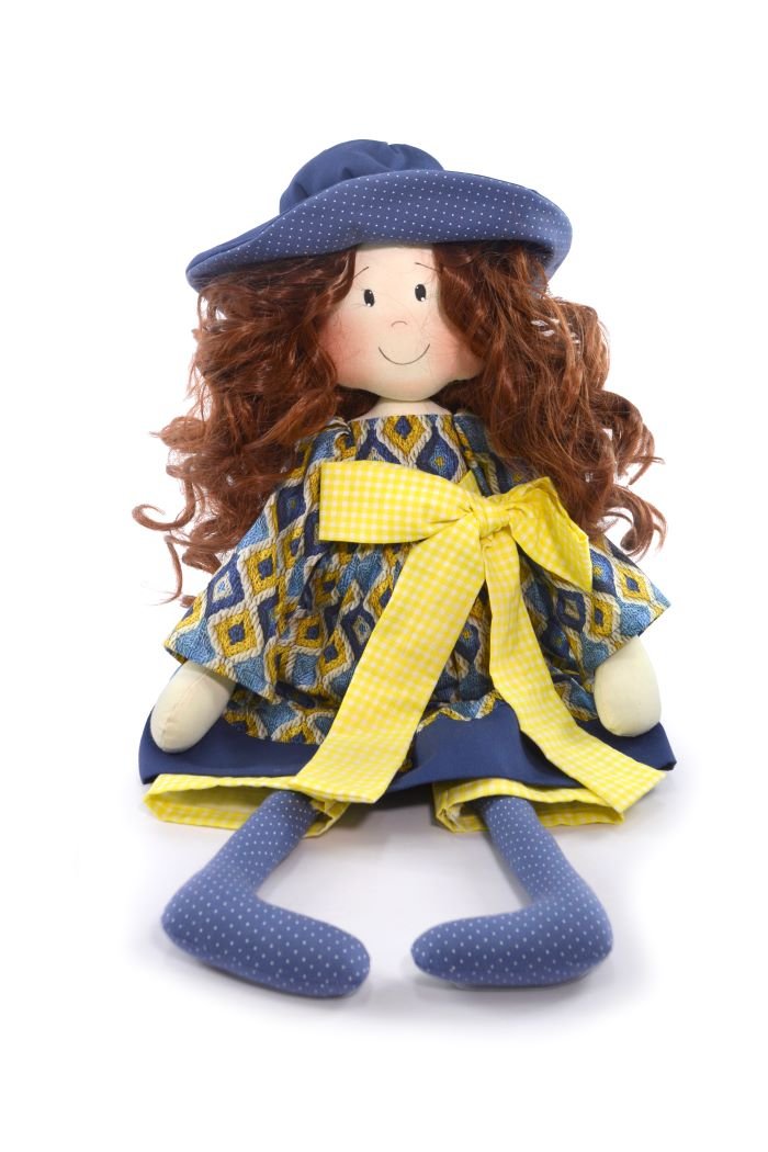 Boneca Amália de costura com vestido estampado em patchwork e chapéu azul, sentada em uma mesa de madeira com materiais de costura ao redor