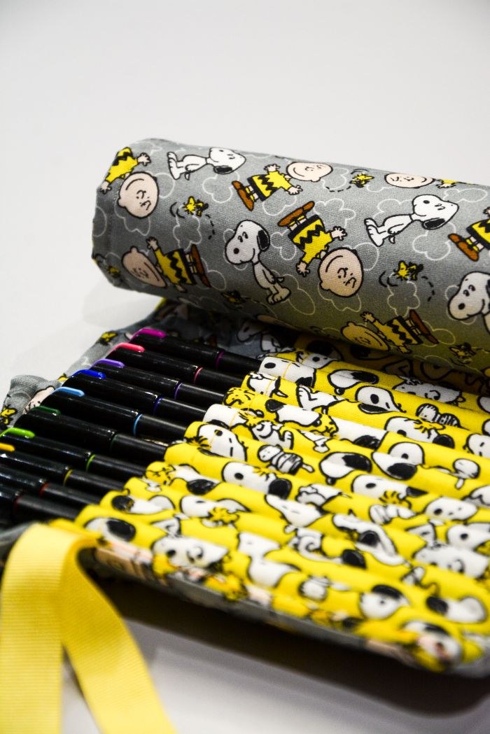 Detalhe do estojo enrolado Snoopy e Charlie aberto com as divisórias em tecido amarelo Peanuts e canetas organizadas
