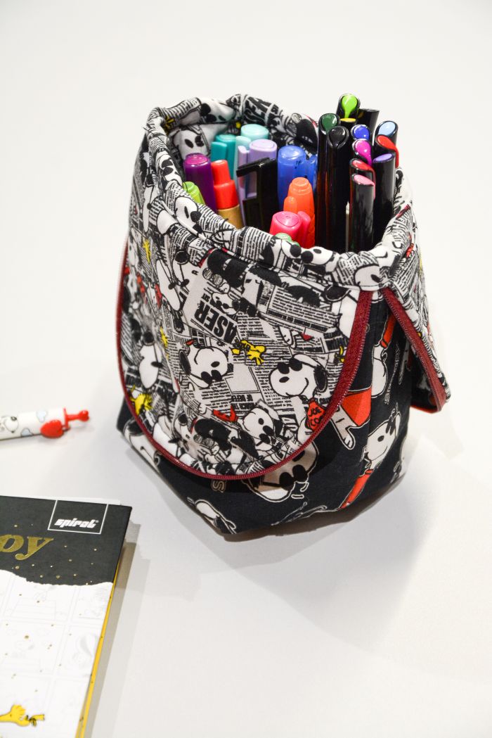 Estojo dobrável Snoopy Cool aberto cheio de canetas coloridas ao lado de estojo fechado – Tecido Peanuts Círculo fundo preto e branco