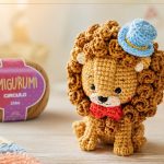 Amigurumi de leão de crochê com chapéu azul, ao lado do novelo Fio Amigurumi Círculo