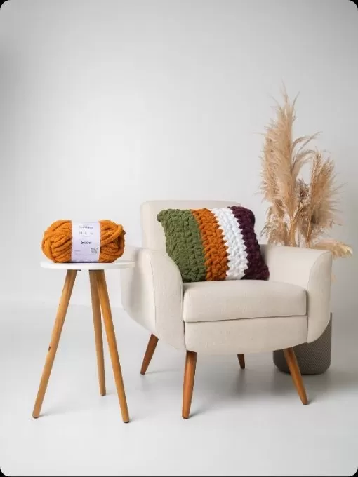 Almofada de crochê com Cisne Hug em listras coloridas na poltrona com novelo laranja ao lado