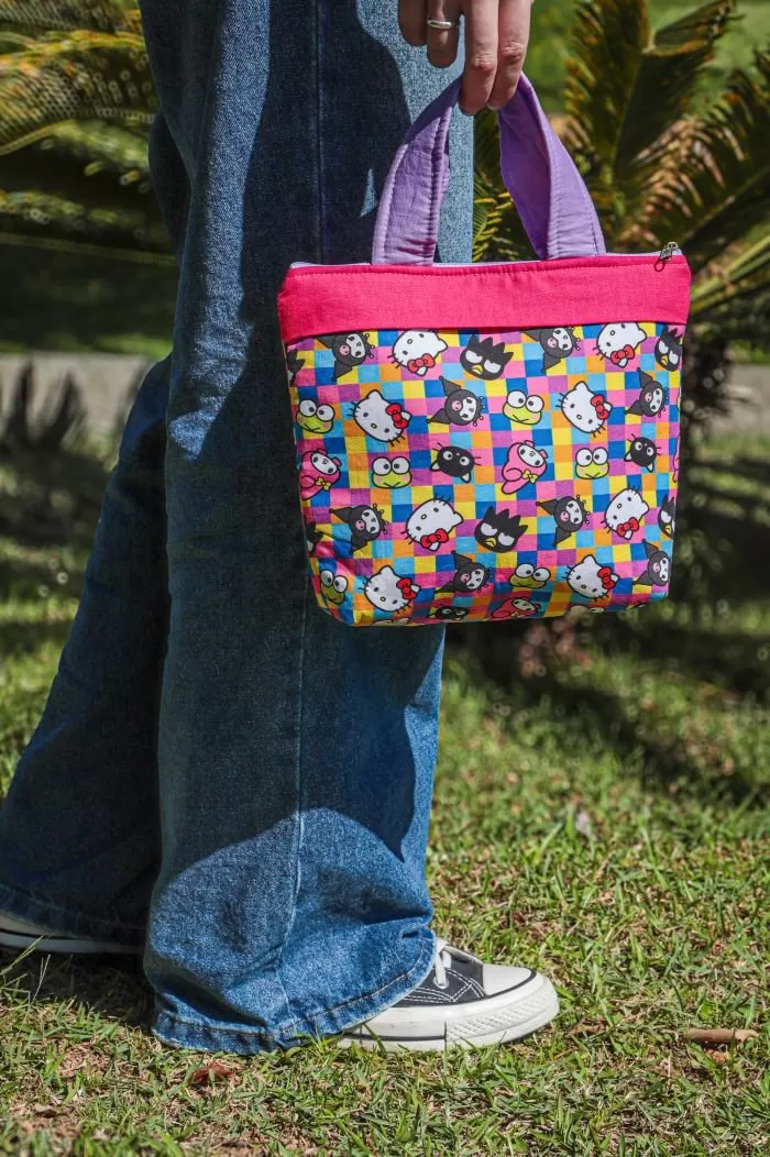 Bolsa Hello Kitty de patchwork com tecido estampado colorido, detalhe em pink e forro lavanda, segurada pela alça