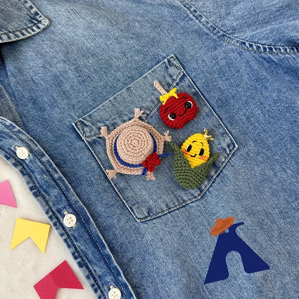Bótons juninos de crochê fixados no bolso de jaqueta jeans — trio chapéu, maçã do amor e milho