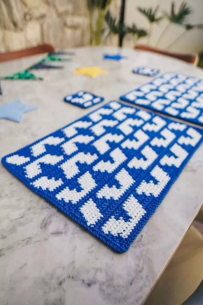 Jogo Americano Copa Copacabana de crochê azul e branco em fio conduzido com padrão de mosaico de Copacabana, sobre mesa com prato e talheres