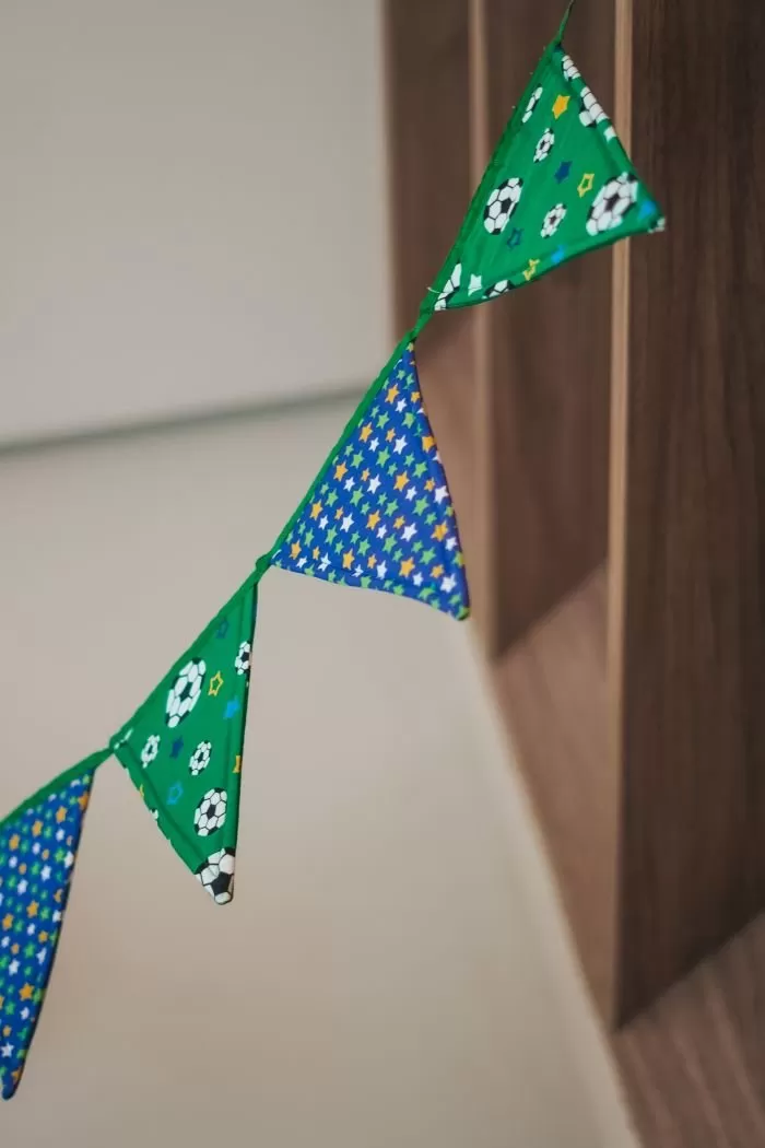 Varal de Bandeiras Copa em patchwork com tecidos verdes e azuis da Coleção Paixão Nacional Círculo, pendurado sobre uma mesa com os tecidos dobrados embaixo