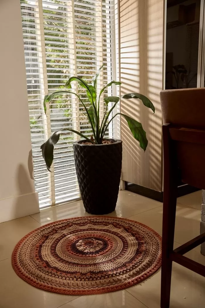 Tapete Ávila de crochê em tons terrosos sobre piso, ao lado de vaso de planta